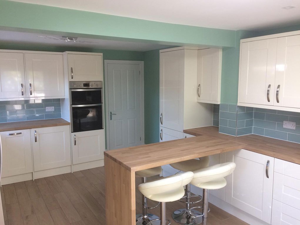 White shaker kitchen with a wood breakfast bar, stools and a sage tiled splashback